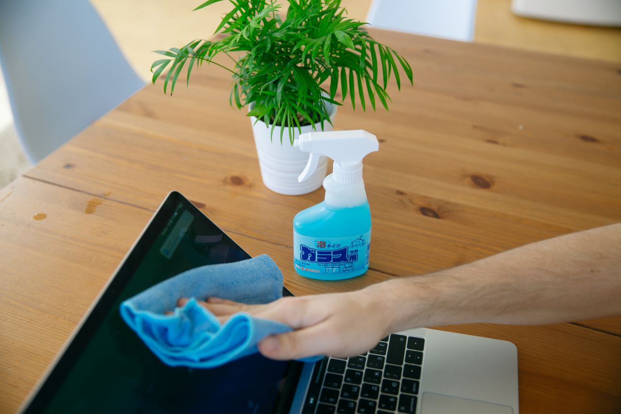 home-img Close-up of hand cleaning a laptop screen with a blue cloth and spray bottle on wooden desk.